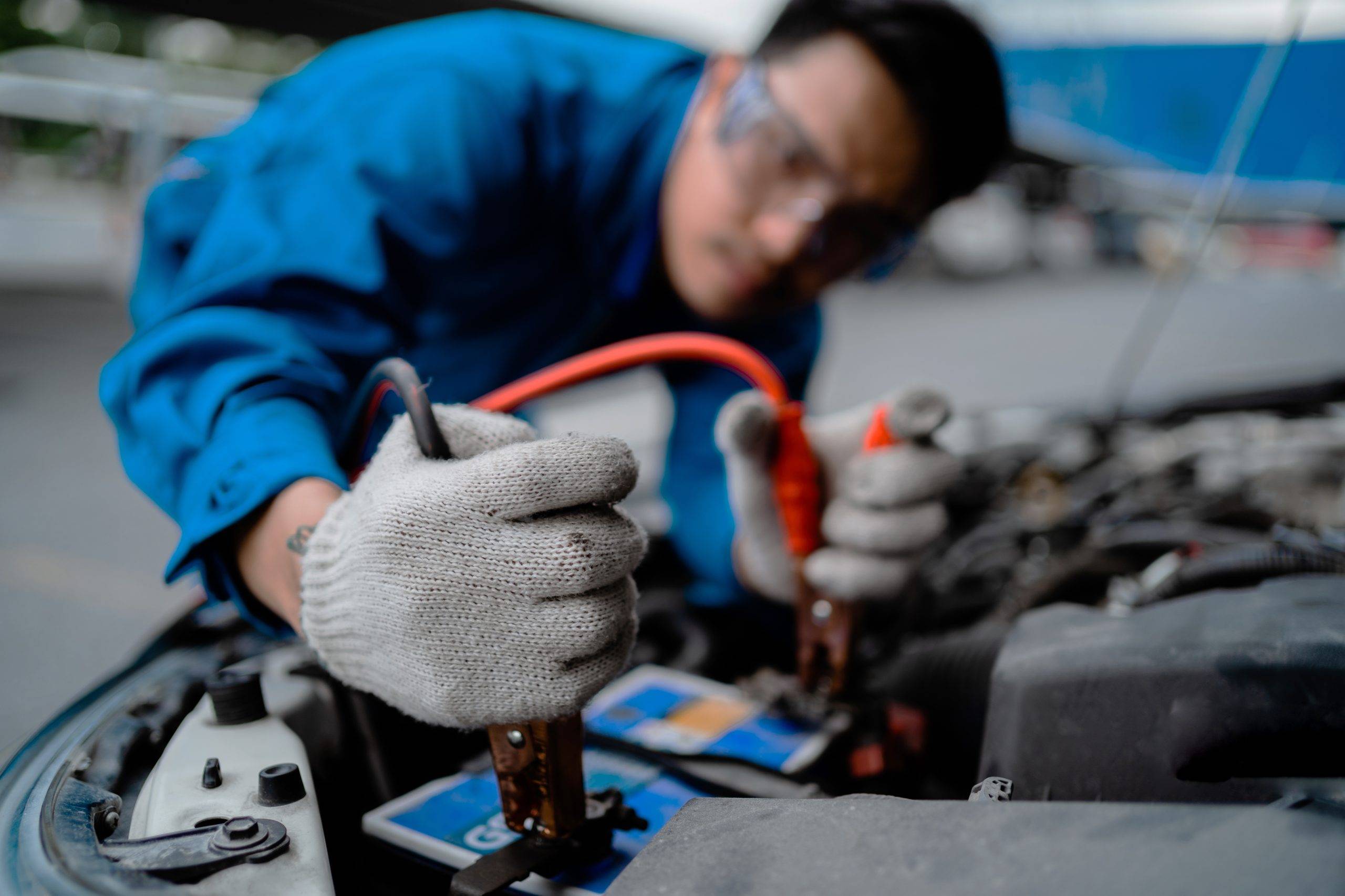 A Man In A Blue Shirt Is Working On A Car Battery. He Is Wearing Gloves And Has A Red Cable In His Hand A Man In A Blue Shirt Is Working On A Car Battery. He Is Wearing Gloves And Has A Red Cable In His Hand