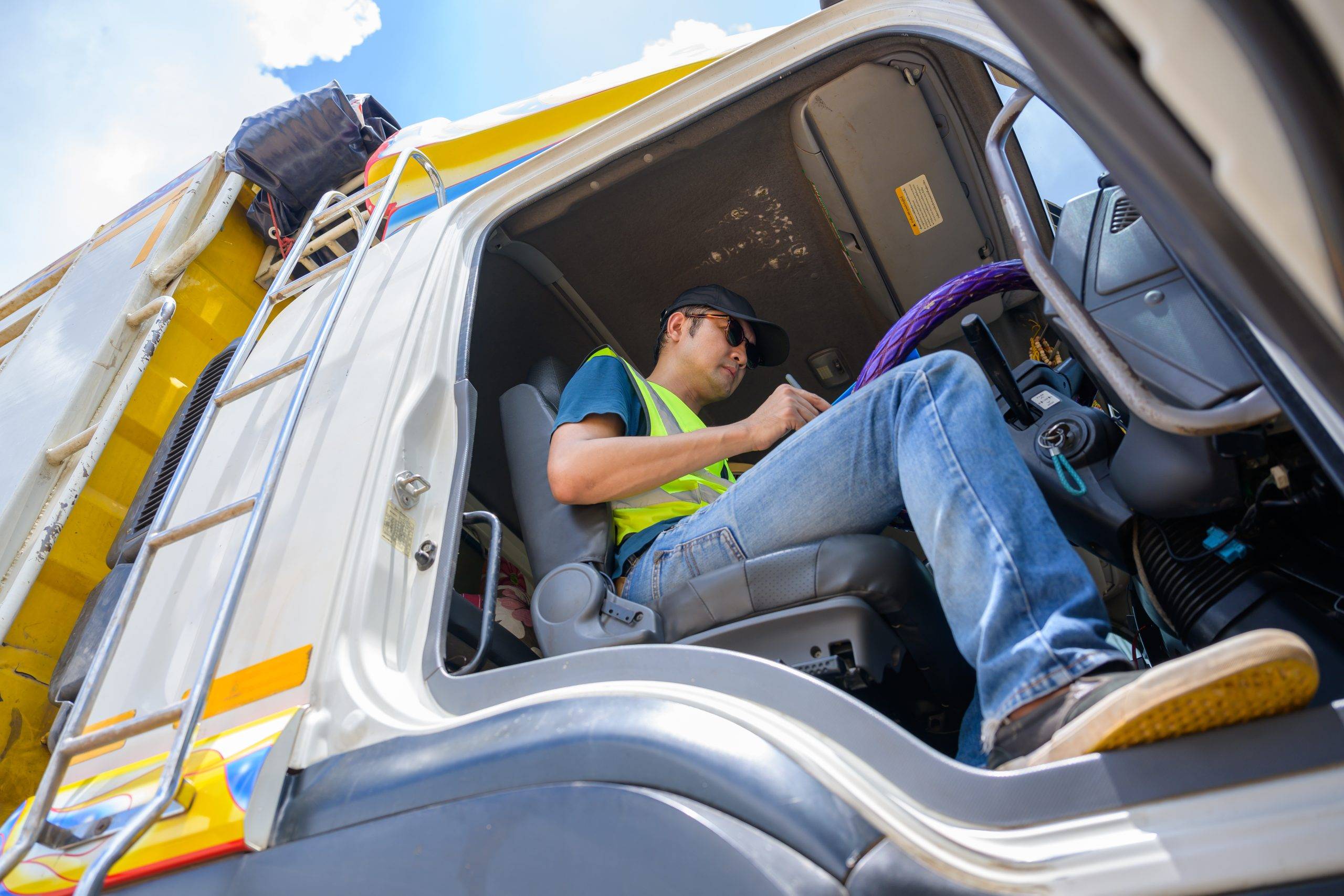 A Truck Driver Wearing A Reflective Vest And Cap Looks At Documents On A Clipboard In The Truck Cabin Of The Transportation Logistics And Trucking Industry. A Truck Driver Wearing A Reflective Vest And Cap Looks At Documents On A Clipboard In The Truck Cabin Of The Transportation Logistics And Trucking Industry.